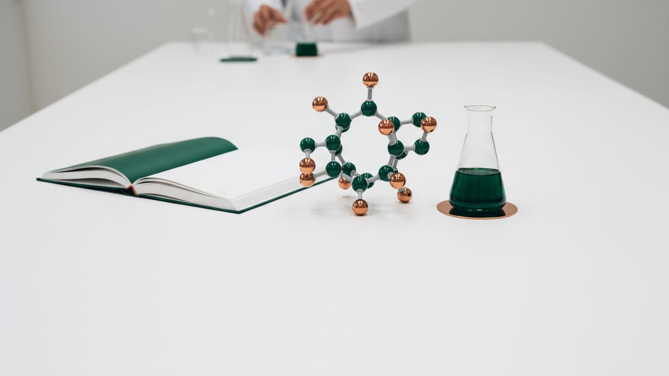Illustration: A wide white laboratory table with a single molecular model, an open blank reference book and a glass flask.
