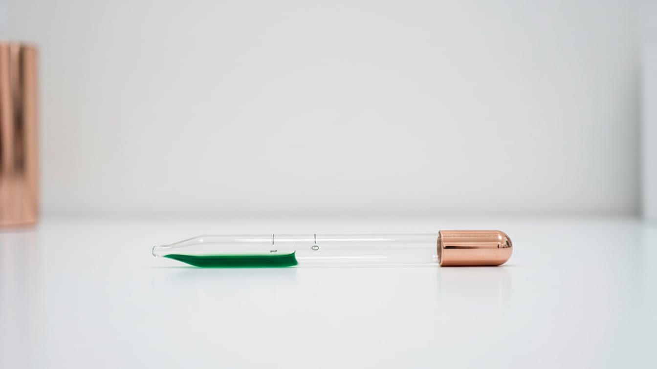 Illustration: A clear glass volumetric pipette with faint green liquid on a white lab bench, copper reflection.