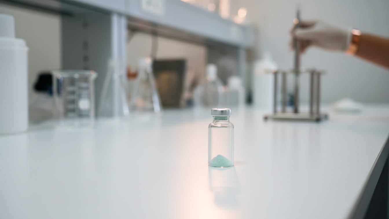 Illustration: A clear glass vial with pale green crystalline powder on a white lab bench with copper reflection.