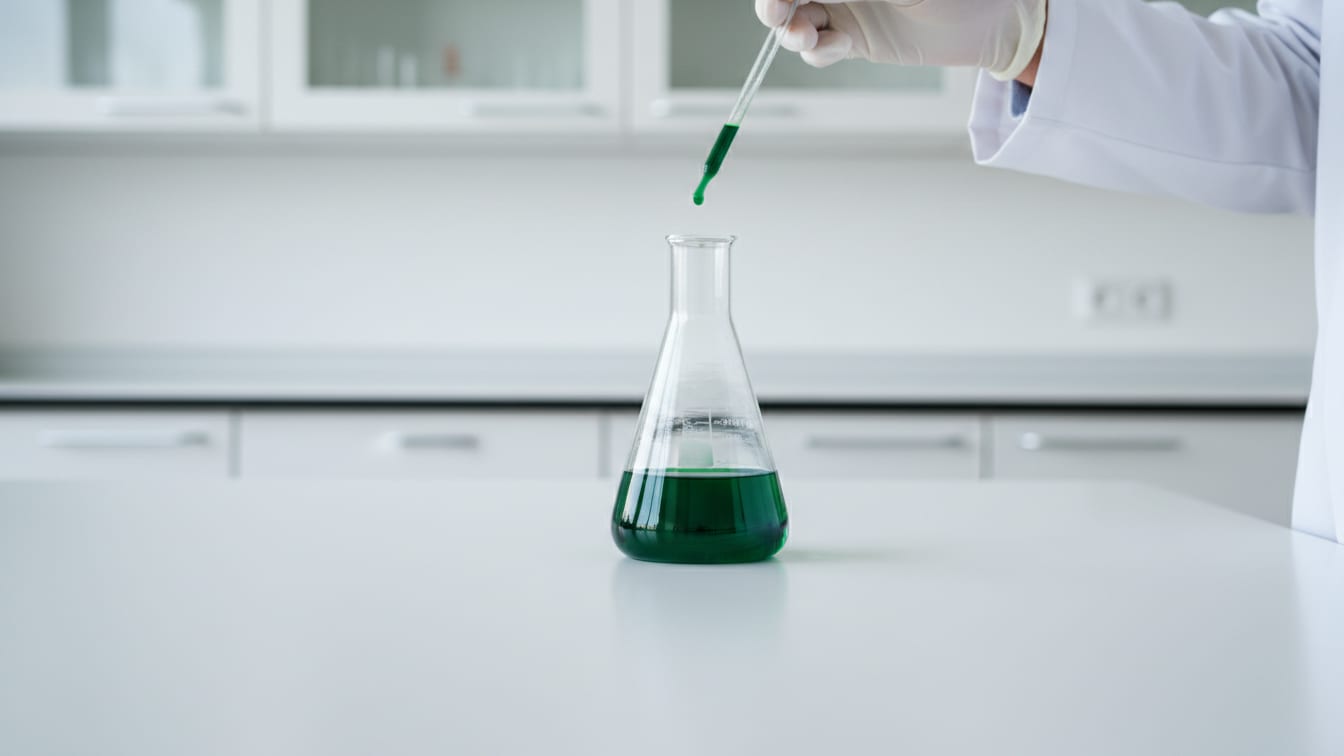 Illustration: A clear glass volumetric flask with faint green liquid on a white lab bench, copper reflections.