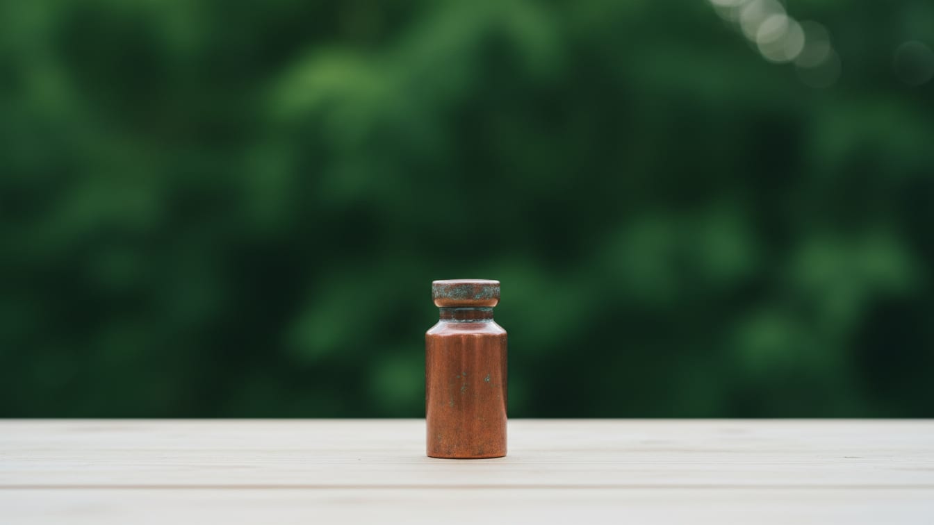 Illustration: An empty, slightly tarnished copper vial on a wooden table with a soft, green background.